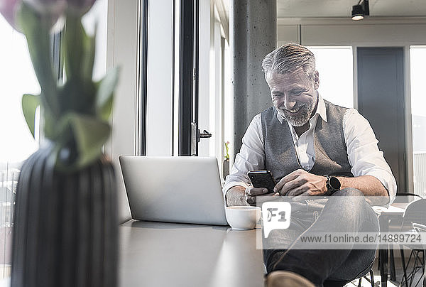 Lächelnder reifer Geschäftsmann mit Laptop und Handy am Fenster sitzend