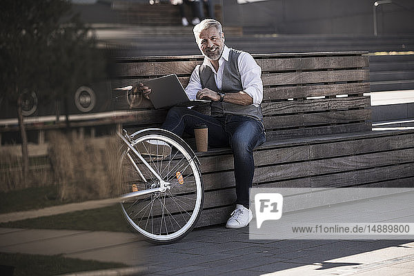 Lächelnder  reifer Geschäftsmann mit Fahrrad  der mit einem Laptop auf einer Bank sitzt