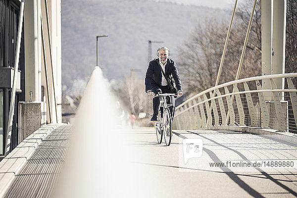 Lächelnder reifer Geschäftsmann mit Kopfhörern beim Fahrradfahren auf einer Brücke in der Stadt