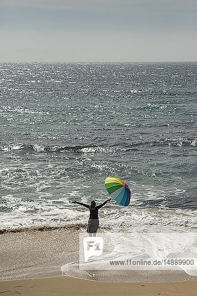 Frau mit buntem Regenschirm am Strand stehend  die Arme erhoben