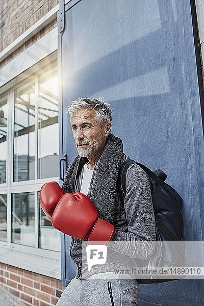 Porträt eines reifen Mannes mit Handtuch  Sporttasche und roten Boxhandschuhen vor der Turnhalle stehend