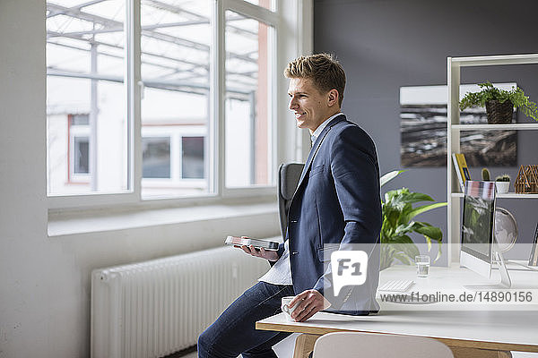 Smiling young businessman sitting on desk in office having a coffee break