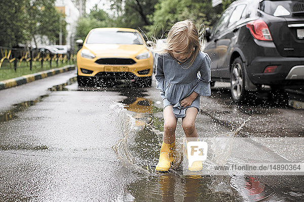 Girl wearing blue dress and rubber boots  jumping in pond on street  yellow car in the background