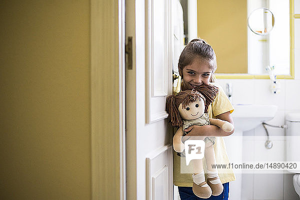 Porträt eines lächelnden kleinen Mädchens  das zu Hause im Türrahmen steht und eine Puppe hält