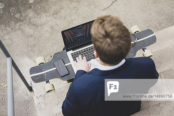 Young businessman with skateboard sitting outdoors on stairs using laptop