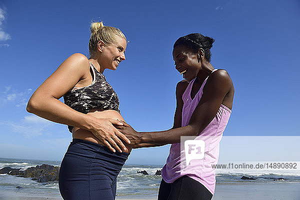 Glückliche Frau berührt den Bauch einer schwangeren Freundin am Strand