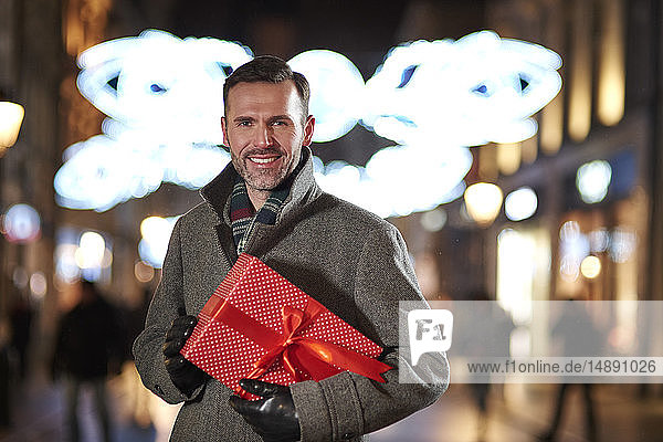 Portrait of smiling man with Christmas present looking at pedestrian area in the evening