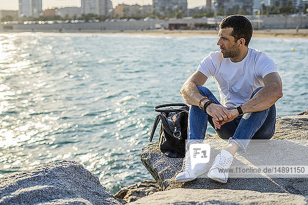 Spanien  Barcelona  Mann sitzt auf einem Felsen vor dem Meer und schaut in die Ferne