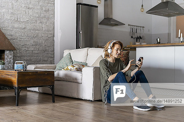Junge Frau sitzt zu Hause auf dem Boden  benutzt ein Smartphone und trägt Kopfhörer