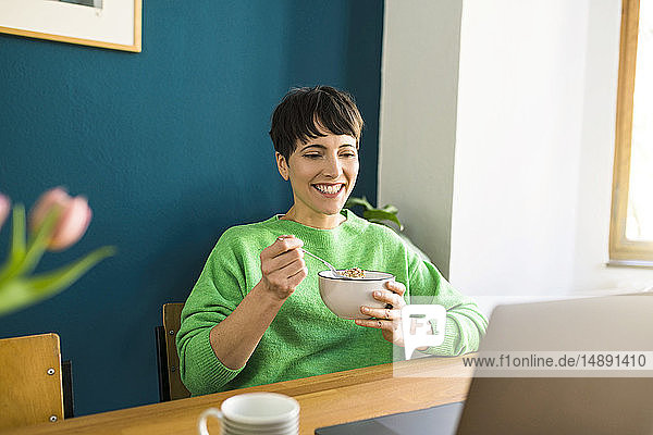 Glückliche kurzhaarige Frau mit grünem Pullover arbeitet mit Laptop im Heimbüro und isst Cornflakes in einer Schüssel