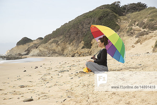 Frau mit buntem Sonnenschirm am Strand sitzend