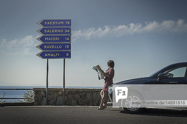 Frau mit Landkarte  die sich an ein Auto mit Straßenschild lehnt