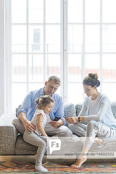 Parents holding smart phones on sofa with their daughter