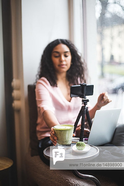 Junge einflussreiche Frau trinkt Matcha-Tee  während sie am Fenster eines Kreativbüros per Handy einen Vlog macht