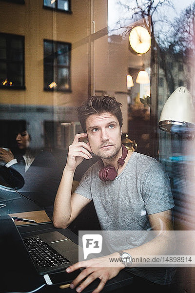Nachdenklicher männlicher Unternehmer sitzt mit Laptop und schaut durch das Fenster im Büro