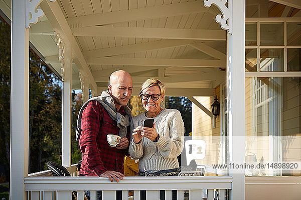Lächelnde reife Frau zeigt kahlköpfigem Freund Mobiltelefon  während sie auf der Veranda steht