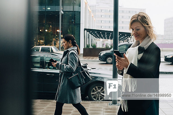 Businesswomen using smart phones while walking on sidewalk in city