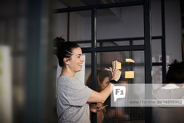 Smiling creative businesswoman sticking adhesive note on glass in office