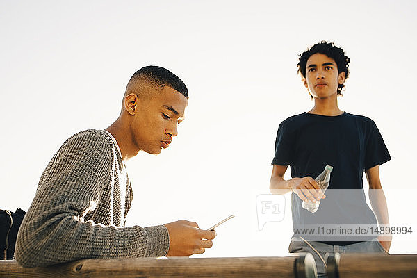 Teenage boy using mobile phone while friend holding water bottle against clear sky during sunny day