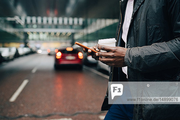 Midsection of businessman with coffee using mobile phone while standing on street in city
