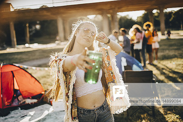 Porträt einer verspielten jungen Frau  die ein Getränk in der Hand hält  während sie auf ein Ereignis an einem sonnigen Tag zeigt