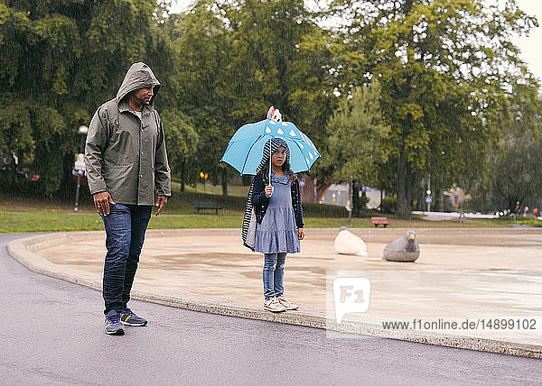 Vater sieht Tochter mit Regenschirm in voller Länge an,  während er bei Regen im Park steht