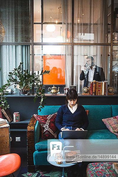 Selbstbewusste Unternehmerin in voller Länge am Laptop auf dem Bürosofa sitzend