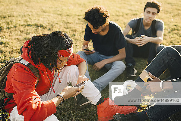 Hochwinkelansicht von Freunden  die Mobiltelefone benutzen  während sie auf dem Feld sitzen
