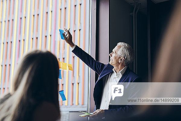 Selbstbewusster reifer Manager klebt während einer Besprechung im Kreativbüro Haftnotizen am Fenster im Sitzungssaal