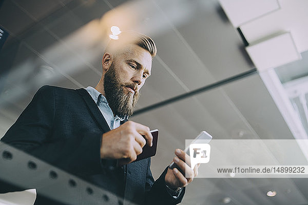Niedrigwinkelansicht eines Geschäftsmannes  der ein Smartphone benutzt  während er seinen Pass am Flughafen in der Hand hält