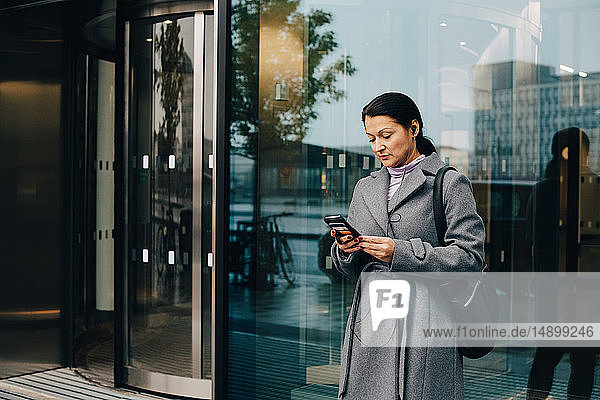 Businesswoman using smart phone while standing against building in city