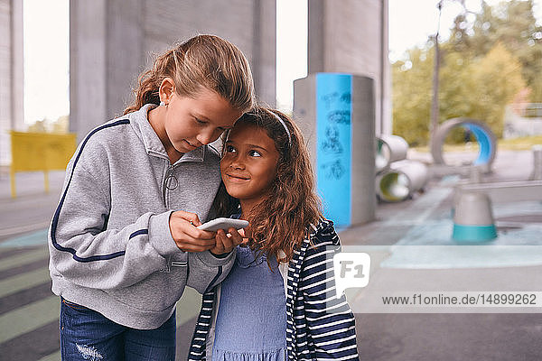Lächelnde Schwestern  die ein Mobiltelefon benutzen  während sie auf dem Fußweg auf dem Spielplatz stehen