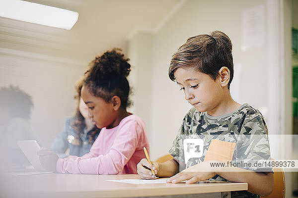 Junge sitzt mit einem Freund zusammen und schreibt am Schreibtisch im Klassenzimmer auf Papier