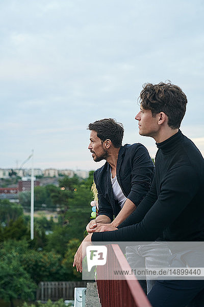 Male friends looking away while standing by railing on terrace against sky