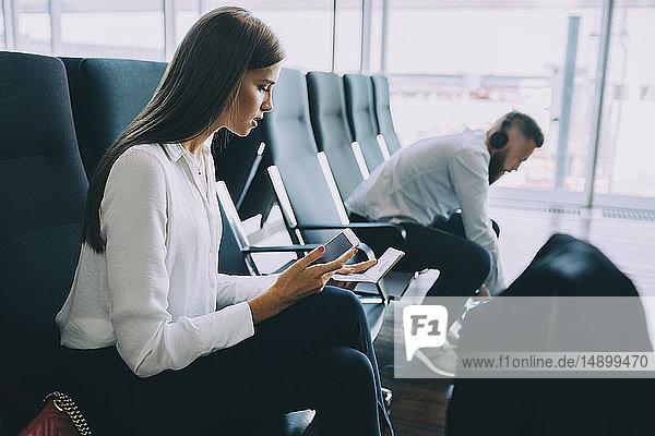 Young businesswoman holding smart phone and passport while sitting at waiting area in airport