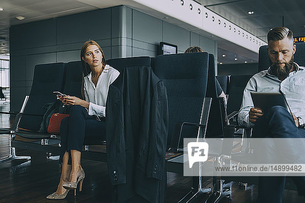 Nachdenkliche junge Geschäftsfrau schaut weg  während sie mit ihrem Kollegen im Abflugbereich des Flughafens sitzt