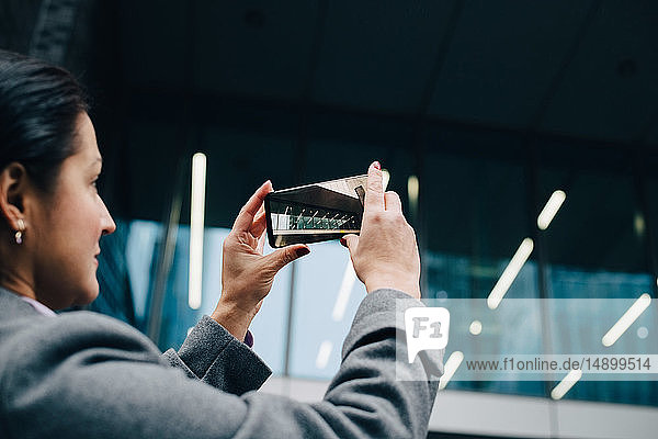 Seitenansicht einer Geschäftsfrau  die ein modernes Gebäude mit einem Smartphone fotografiert  während sie in der Stadt steht