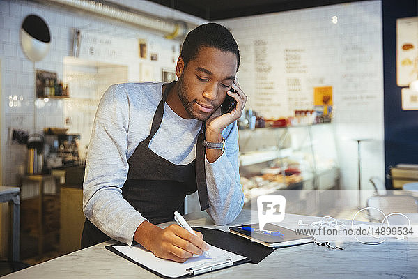 Selbstbewusster junger männlicher Angestellter spricht durch ein Smartphone  während er in einem Feinkostladen an der Zwischenablage schreibt