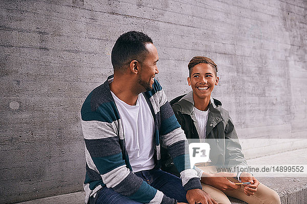 Fröhliche Väter und Söhne schauen einander von Angesicht zu Angesicht an  während sie auf dem Spielplatz an der Wand sitzen