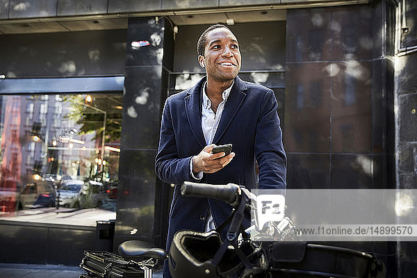 Lächelnder junger männlicher Pendler  der ein Smartphone benutzt  während er mit einem Elektrofahrrad gegen ein Gebäude in der Stadt steht