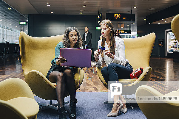 Multi-ethnische Kolleginnen diskutieren in voller Länge über Laptop und Kreditkarte  während sie in der Flughafen-Lobby sitzen
