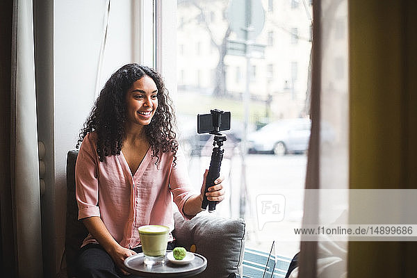 Selbstbewusste junge einflussreiche Frau  die am Fenster sitzend am Fenster eines kreativen Büros durch ein Mobiltelefon auf einem Stativ vloggt