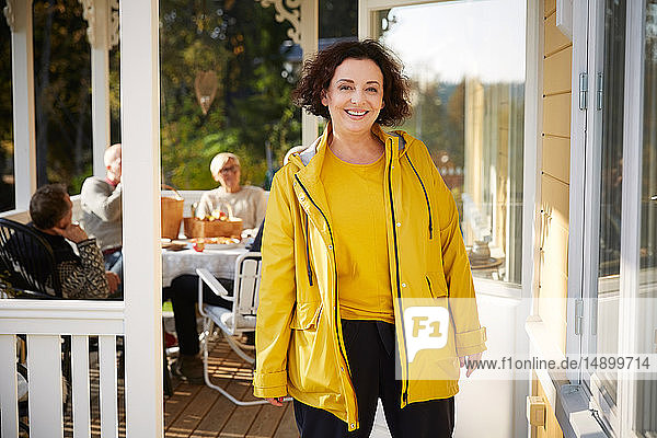 Porträt einer fröhlichen  reifen Frau in gelber Jacke  die auf der Veranda steht  während im Hintergrund Freunde sitzen