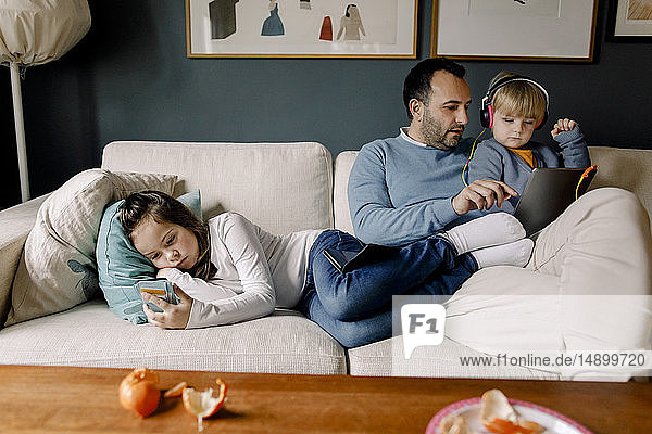 Girl using mobile phone while father showing digital tablet to sister on couch in living room at home