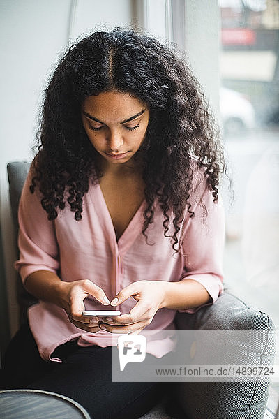 Junge Geschäftsfrau  die im Kreativbüro am Fenster sitzt und Textnachrichten von einem Smartphone aus verschickt