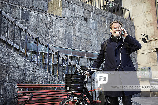 Smiling man talking on smart phone by electric bicycle against building