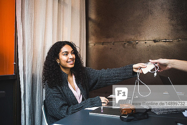 Lächelnde junge berufstätige Frau übergibt Ladegerät an männlichen Kollegen am Schreibtisch im Büro