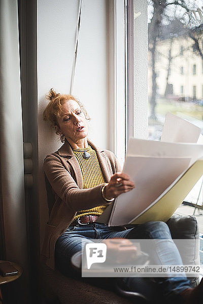 Mature female illustrator looking at papers on window sill in office
