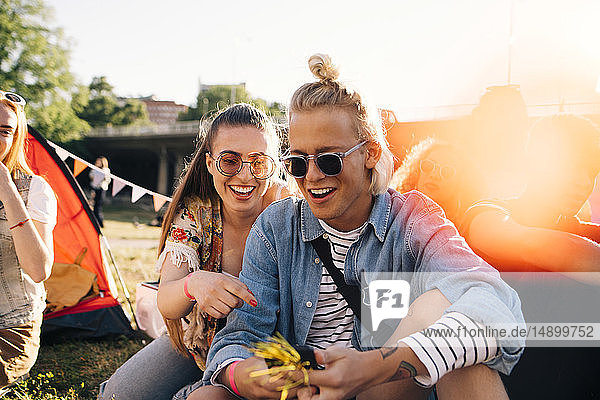 Lächelnde männliche und weibliche Freunde schauen auf das Smartphone  während sie an einem sonnigen Tag auf dem Rasen sitzen