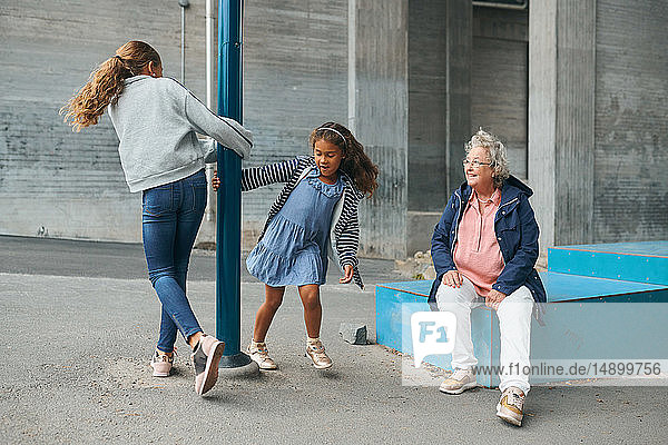 Smiling grandmother looking at playful granddaughters spinning around pole at playground